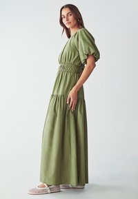 Abito maxi in tessuto verde oliva con maniche a sbuffo, vita arricciata e gonna a strati. Abbinato a ballerine in rete bianche.