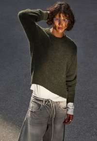 Olivgrüner Strickpullover mit legerem Schnitt und verkürztem Saum, kombiniert mit hellgrauen Jeans mit Kordelzug und vorderen Taschen.