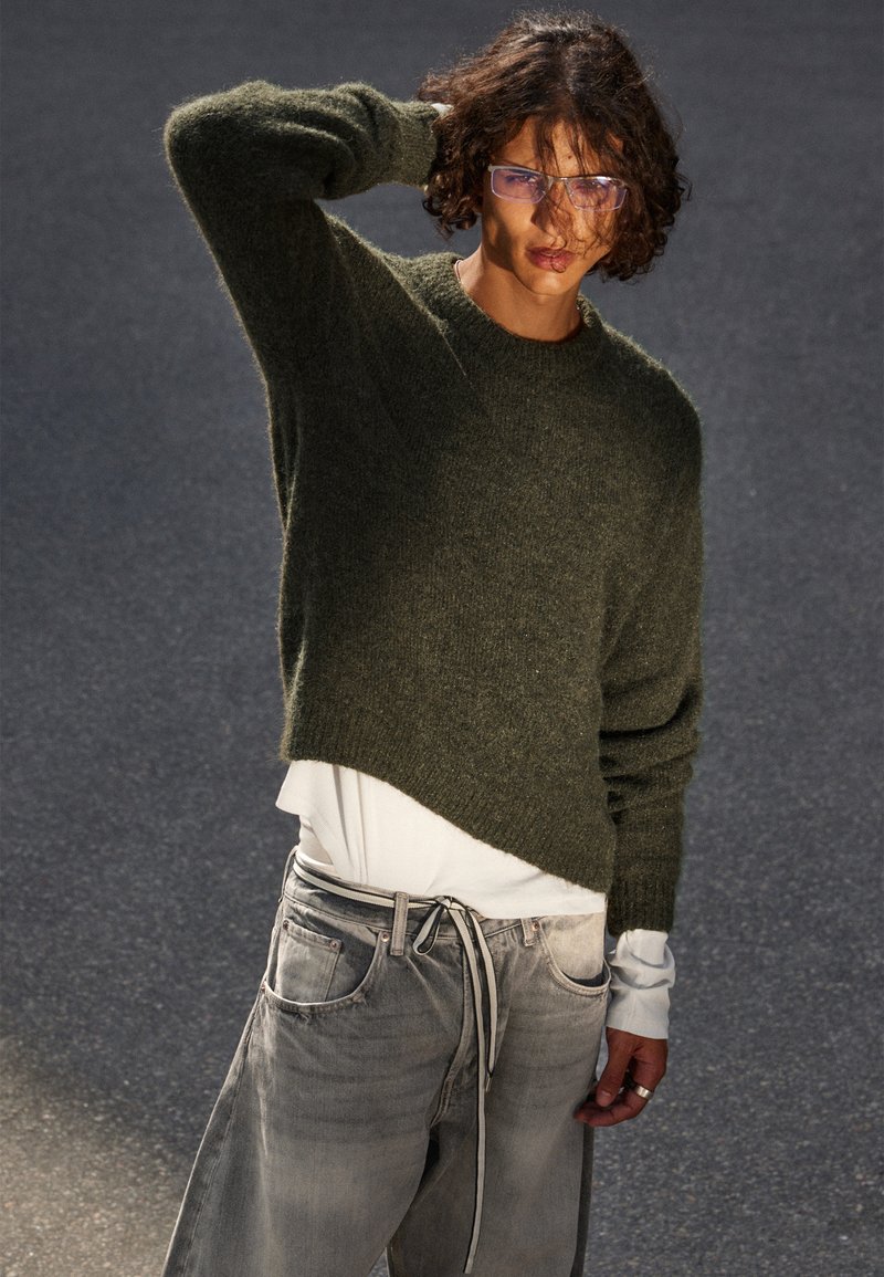 Olivgrüner Strickpullover mit legerem Schnitt und verkürztem Saum, kombiniert mit hellgrauen Jeans mit Kordelzug und vorderen Taschen.