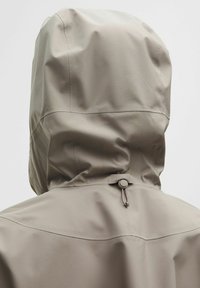 Ljusbeige vattentät jacka med en åtsittande huva, elastisk dragsko och slät yta. Sömnadsdetaljer på baksidan.