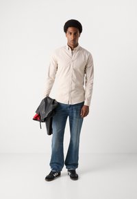 Ανοιχτό καφέ πουκάμισο, τζιν παντελόνι, μαύρα αθλητικά παπούτσια με λευκές ρίγες, κρατώντας μία μαύρη δερμάτινη ζακέτα με κόκκινη επένδυση.