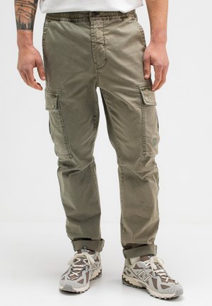 Cargohosen aus olivgrünem Stoff mit einem elastischen Bund, Seitentaschen und gesteppten Details. Tragen mit grauen und beigen Sportschuhen.