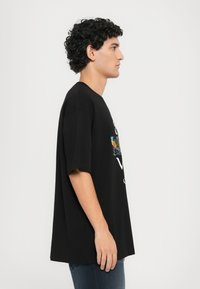 Black oversized t-shirt in cotton, featuring a graphic design in color and white text at the front. Side view shows loose fit and short sleeves.