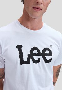 Mann iført hvit t-skjorte med stor svart "Lee"-logo, tatovering synlig på venstre overarm, nøytral grå bakgrunn.