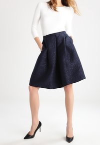 Marineblaue A-Linien-Rock mit strukturierter Stoffqualität, Seitentaschen und tailliertem Sitz, kombiniert mit einem weißen Langarmshirt und schwarzen High Heels.