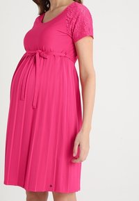 Rosa Schwangerschaftskleid mit kurzen Spitzenärmeln, tailliertem Bindegürtel und plissiertem Rock. Weicher Stoff, knielanges Design.