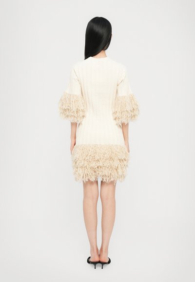 Robe en tricot crème avec une texture côtelée, dotée de manches surdimensionnées et d'un ourlet avec franges. Coupe ajustée, longueur mi-cuisse, associée à des chaussures noires.