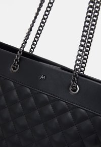 Sac cabas noir en similicuir matelassé avec poignées doubles en chaîne et un logo discret. Présente une texture lisse et un design élégant.