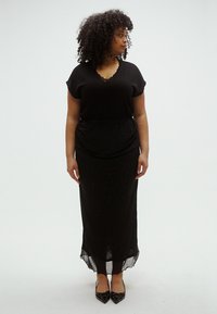 Svart kjole med en helfarget overdel med blonder ved halsen og et gjennomsiktig skjørt med paljetter, kombinert med spisse svarte hæler.