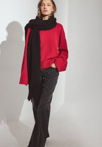 Suéter rojo de punto con puños y dobladillo acanalados, combinado con una larga bufanda negra, llevado sobre pantalones negros de pierna ancha. Textura suave, ajuste casual.