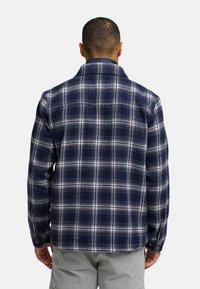 Chemise en flanelle à carreaux bleu marine et blanc, avec un col évasé, des manches longues et des poignets à bouton, présentant une texture douce et un ourlet droit.