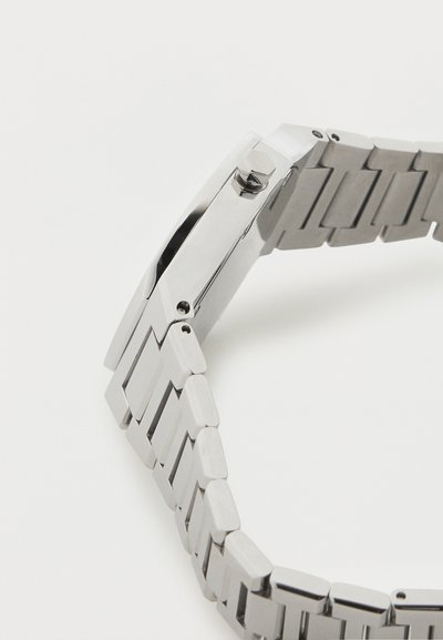 Ασημένιο μεταλλικό λουράκι ρολογιού με συνδεδεμένα ορθογώνια τμήματα και κορώνα στην πλευρά της θήκης του ρολογιού, σε γωνία πάνω σε λευκό φόντο.