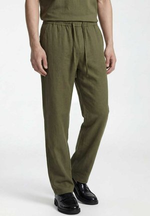 Man draagt olijfgroene broek met trekkoord en zwarte leren loafers, staande tegen een effen witte achtergrond.