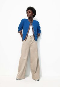 Cardigan texturé bleu sur une chemise à carreaux, un haut blanc et un pantalon large beige. Des baskets grises complètent l'ensemble. Fond blanc uni.
