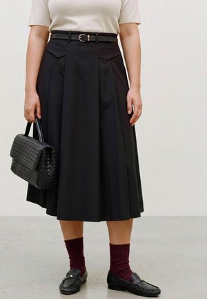 Une personne porte une jupe midi noire plissée avec une ceinture, un sac à main noir tressé, des chaussettes bordeaux et des mocassins noirs.