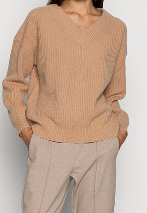 Beżowy sweter dzianinowy z dekoltem w serek, prążkowaną teksturą i długimi rękawami, w połączeniu z jasnobeżowymi spodniami. Posiada luźny krój.