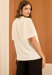 Weiße Bluse mit Punktstruktur, kurzen Ärmeln und Spitzenbesatz am Kragen. Kombiniert mit schwarzen Hosen, vor einem Holz-Hintergrund präsentiert.