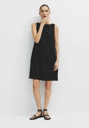 Žena stojaca pri bielej stene, oblečená v čiernych bezrukávoch a čiernych sandáloch na plochom podpätku, s jednou rukou pri ústach.