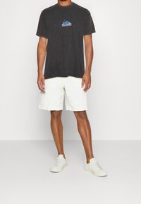 Černé bavlněné tričko s malou barevnou grafikou na hrudi, kombinované s bílými bavlněnými šortkami a béžovými teniskami.