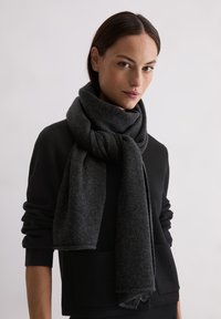 Vrouw met donker haar die een zwarte trui en een dikke, donkergrijze gebreide sjaal om haar nek draagt.