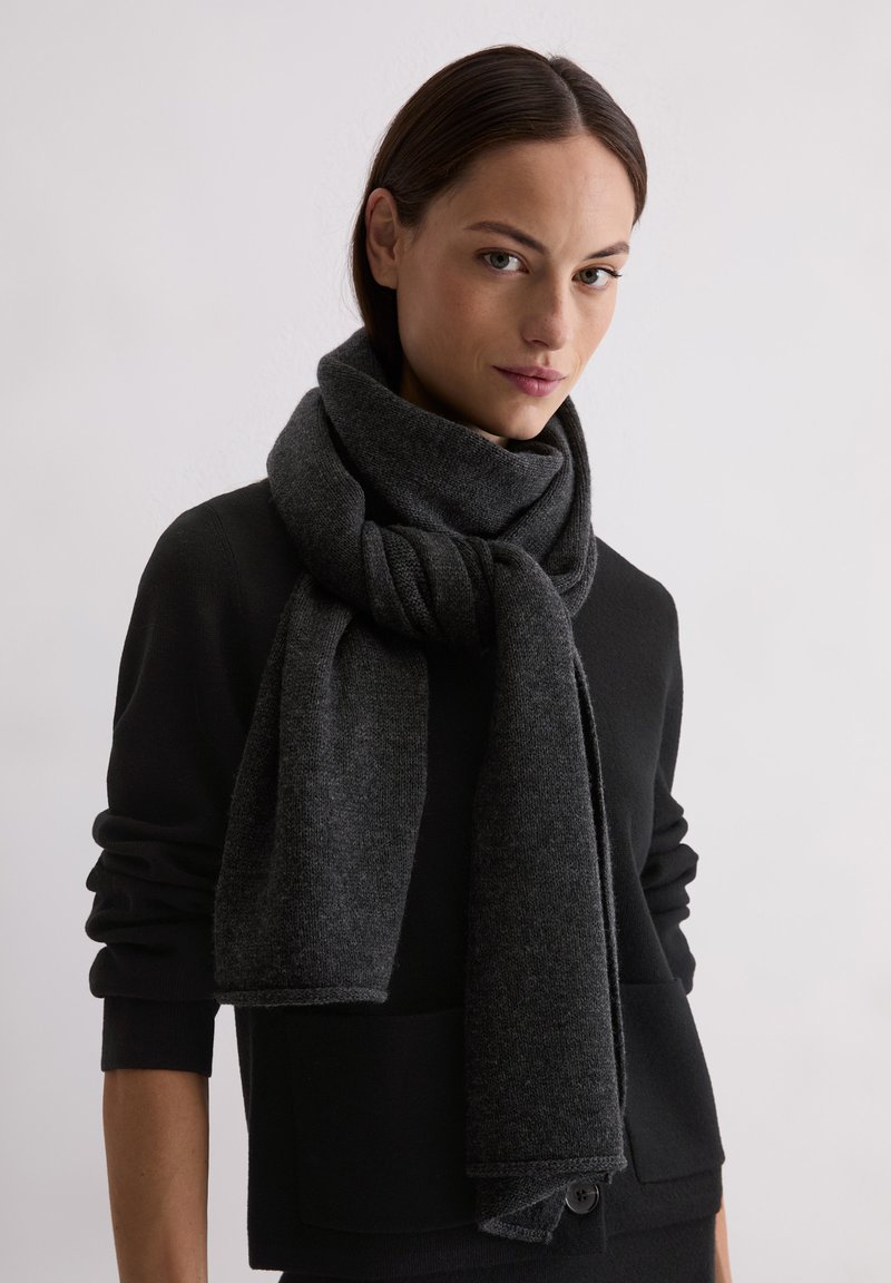 Vrouw met donker haar die een zwarte trui en een dikke, donkergrijze gebreide sjaal om haar nek draagt.