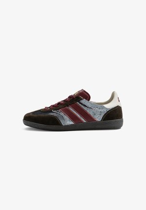 Zapatilla deportiva de perfil bajo con puntera y talón de ante marrón oscuro, paneles laterales con lentejuelas plateadas y rayas y cordones burdeos sobre fondo blanco.