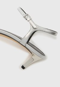 Ασημένιο μεταλλικό σανδάλι με λουράκια, μέτριο τακούνι, ρυθμιζόμενο πόρπη και λεπτή υφή. Διαθέτει κομψό σχεδιασμό και μαύρη μύτη τακουνιού.