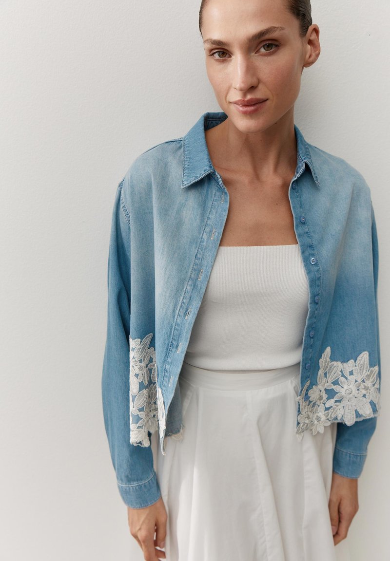 Woman wearing a light blue denim jacket with white floral lace trim over a white tube top and white skirt, standing against a plain wall.