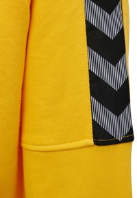 Gul sweatshirt med texturerad tyg, som har en svart sidopanel med ett sicksackmönster i vita ränder och en kontrasterande sömdetalj.