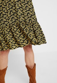 Svart blommig klänning med mönster av gula blad, volangkant, kombinerad med knä-långa bruna mocka cowboyboots. Enkel design och struktur.