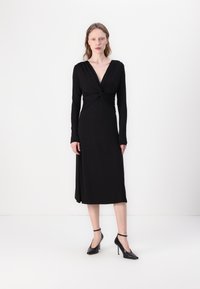Black long-sleeve dress with a deep V-neck and twisted front detail. Made of smooth fabric. Paired with black high-heeled shoes.