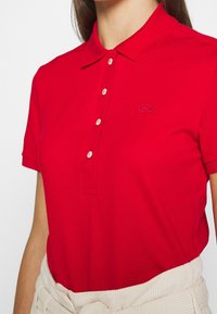 Rød polo skjorte med ribbet krave, knaplukning og Lacoste logo broderet på venstre bryst. Korte ærmer og tekstureret stof.