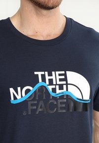 Námořnická modrá tričko vyrobené z bavlny, se grafickým designem bílého a modrého nápisu "THE NORTH FACE" a modrým vzorem vln.