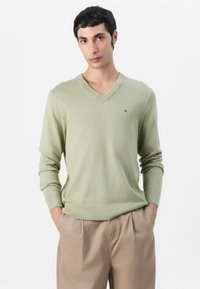 Lys grønn V-hals genser laget av myk stoff. Den har lange ermer og ribbede mansjetter. Liten logo på venstre bryst. Kombinert med beige bukser.