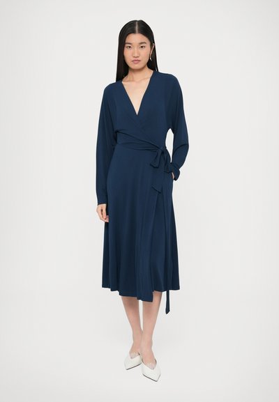 Tumši zila wrap kleita ar gariem piedurknēm, dziļu V veida kakla izgriezumu un sasienamu vidukli. Gluda auduma un plūstoša svārku daļa, saskaņota ar baltiem, smailiem apaviem.