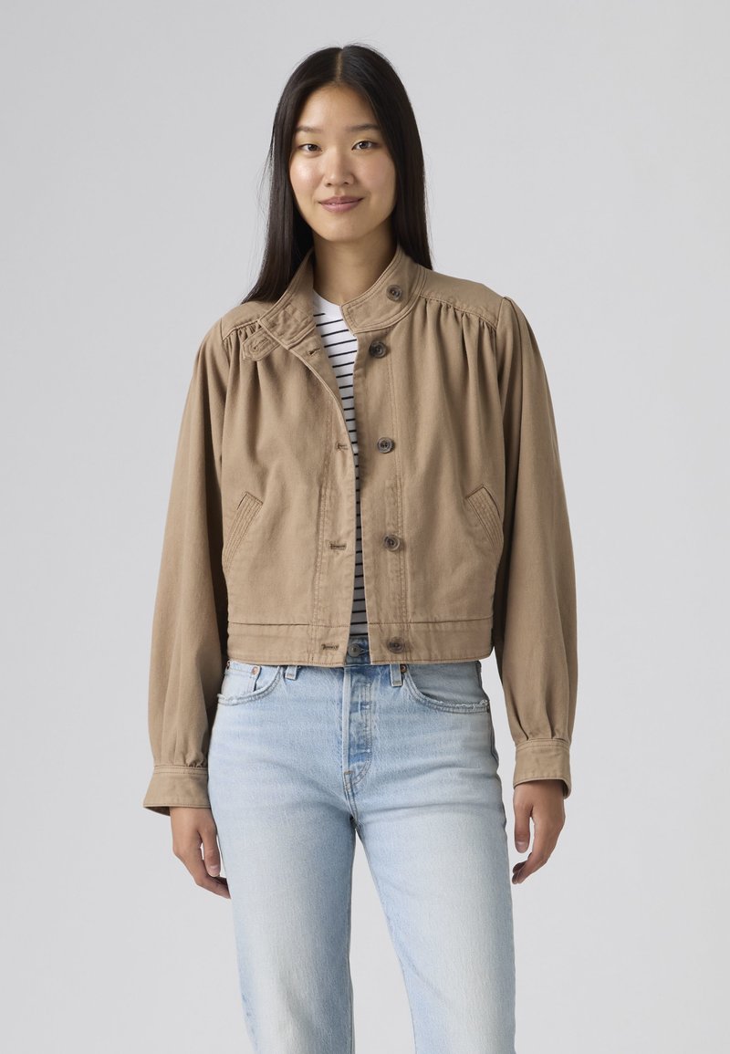 Mujer joven con cabello lacio y oscuro, vestida con una chaqueta beige abotonada sobre una camisa de rayas y jeans azul claro, posando frente a un fondo liso.
