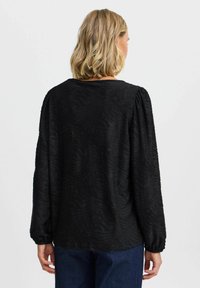 Schwarze Bluse mit strukturierten, wellenförmigen Mustern, langen Ärmeln und rundem Ausschnitt. Der Stoff wirkt leicht und softly strukturiert.
