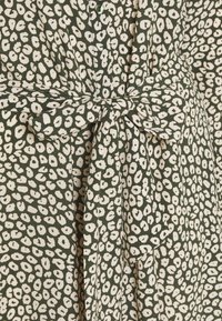 Abito verde con macchie bianche irregolari, caratterizzato da una vita annodata e dettagli con bottoni. Realizzato in tessuto leggero con una consistenza morbida.