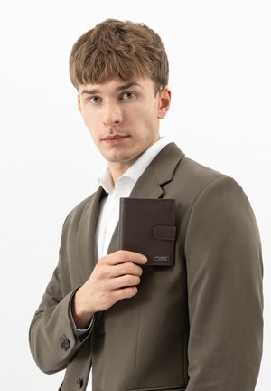 Junger Mann im grauen Anzug hält eine geschlossene dunkelbraune Brieftasche nahe der Brust und schaut direkt voraus vor einem schlichten weißen Hintergrund.