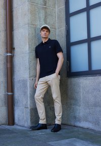 Must vald musta polo-särk, millel on väike logo, beežid chinos-püksid ja mustad pahkluu saapad. Mudel seisab kiviseina vastas, kandes heledat mütsi.