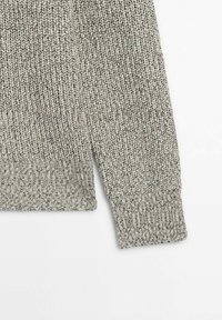 Gros plan sur la manche d'un pull gris en maille avec un poignet côtelé et un motif de points texturés sur fond blanc.