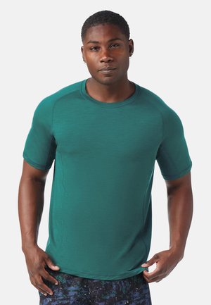 Uomo che indossa una maglietta sportiva a maniche corte color turchese e pantaloncini scuri con motivo, in piedi davanti a uno sfondo bianco semplice.