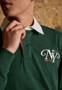 Grön polo-skjorta med vit krage, med broderad "NY" logotyp och texten "NEW YORK". Materialet verkar mjukt med ribbad textur.