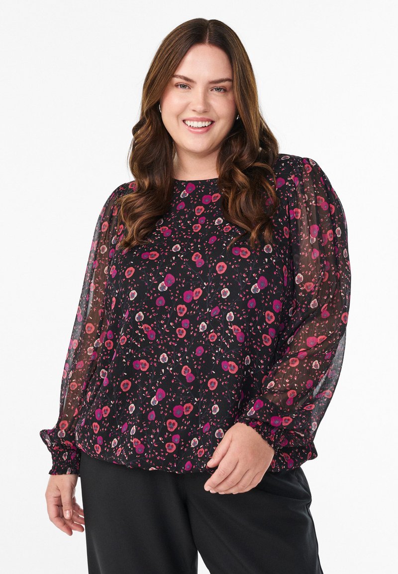Lächelnde Frau mit langen, welligen braunen Haaren trägt eine schwarze Bluse mit transparenten Puffärmeln und pinkem Blumenmuster, kombiniert mit schwarzen Hosen.