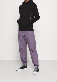 Hombre con sudadera negra con capucha y bolsillos frontales, pantalones cargo morados y zapatillas negras de caña alta, de pie frente a un fondo blanco liso.