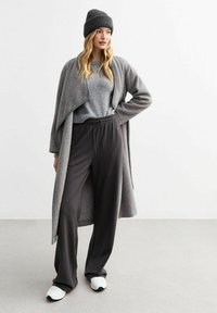 Grå stickad mössa, mjuk grå tröja och en lång grå cardigan, kombinerat med lösa svarta byxor och vita sneakers. Släta material och avslappnad passform.