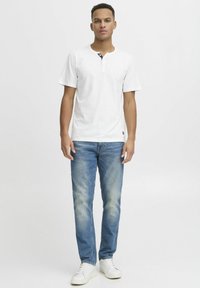 Cămașă henley albă cu mânecă scurtă și gât rotund, cu accent de nasture bleumarin, asociată cu blugi de denim albaștri deschiși și adidași albi.