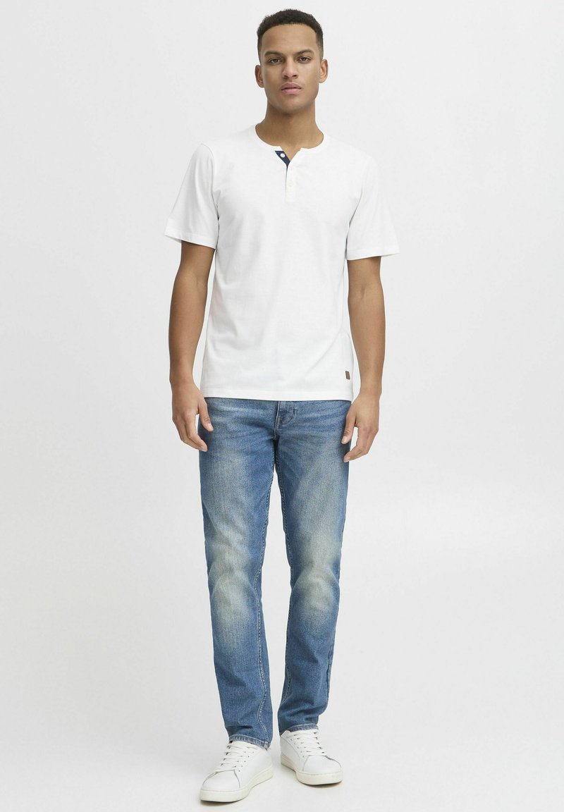 Cămașă henley albă cu mânecă scurtă și gât rotund, cu accent de nasture bleumarin, asociată cu blugi de denim albaștri deschiși și adidași albi.