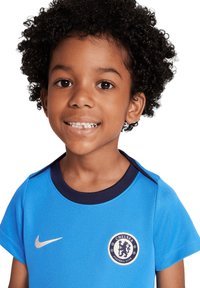 Bambino sorridente con capelli ricci che indossa una maglia del Chelsea Football Club blu con il logo Nike su uno sfondo bianco.
