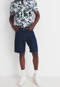 Camicia bianca a maniche corte con motivo floreale, abbinata a pantaloni shorts di denim scuri e sneakers bianche. La camicia presenta un design a bottoni.